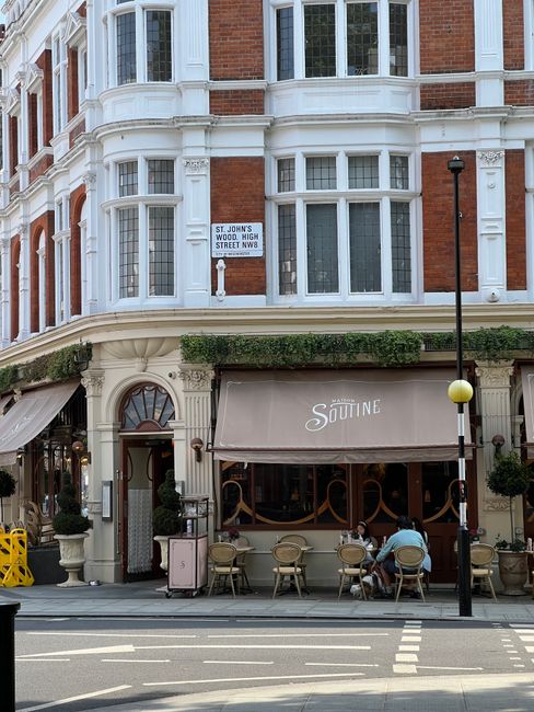 A street view of a historic red brick building with white decorative trim and large bay windows above an outdoor seating area of a café named 'Soutine.' The café has a beige awning, with several emp